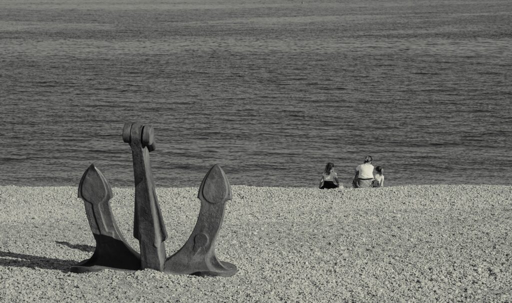 Black and white photo of a family sitting by the sea with a large anchor on a pebbly beach.